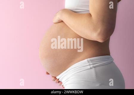 Descrizione: Sezione centrale di una madre in piedi irriconoscibile che tiene delicatamente la pancia incinta molto rotonda. Vista laterale. Sfondo rosa. Colpo luminoso. Foto Stock