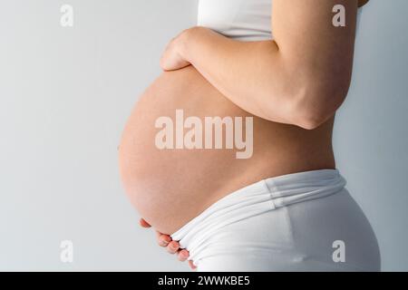 Descrizione: Parte centrale di una donna in piedi irriconoscibile che tiene delicatamente il suo bordino incinta molto rotondo. Vista laterale. Sfondo bianco. Colpo luminoso. Foto Stock