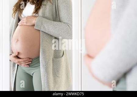 Descrizione: Vista laterale della sezione centrale di una donna irriconoscibile che tiene delicatamente la pancia davanti a uno specchio. Gravidanza primo trimestre - settimana 18. Foto Stock