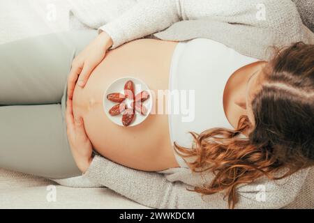 Descrizione: Vista dall'alto di una donna seduta sul divano che mangia Daddles mentre tiene delicatamente la pancia negli ultimi mesi di gravidanza. Gravidanza terzo tr Foto Stock