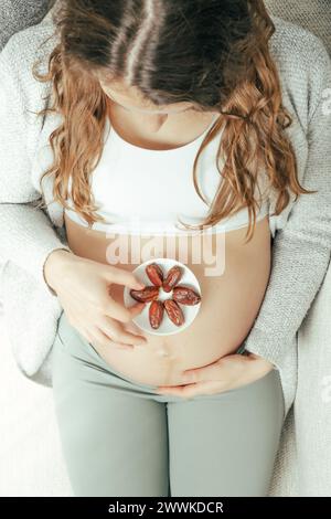 Descrizione: Vista dall'alto di una donna seduta sul divano che mangia Daddles mentre tiene delicatamente la pancia negli ultimi mesi di gravidanza. Gravidanza terzo tr Foto Stock