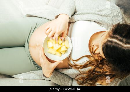Descrizione: Vista dall'alto di una donna seduta sul divano che mangia pezzi di Ananas mentre tiene delicatamente la pancia negli ultimi mesi di gravidanza. Gravidanza Foto Stock