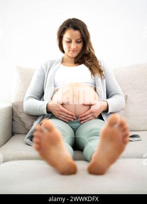 Descrizione: Vista frontale di una donna sorridente seduta su un divano che tiene orgogliosamente la pancia in attesa del bambino durante l'ultima fase della gravidanza. Foto Stock