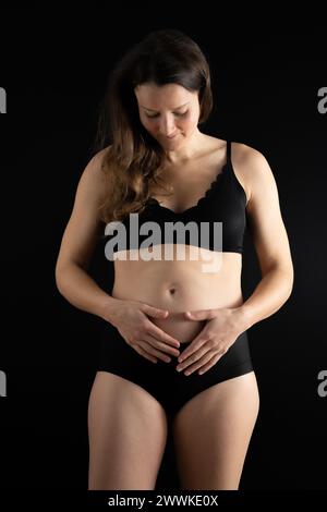 Descrizione: Vista di una donna felice che tiene dolcemente la pancia durante i primi mesi di gravidanza. Gravidanza primo trimestre - settimana 18. Vista frontale. bac nero Foto Stock