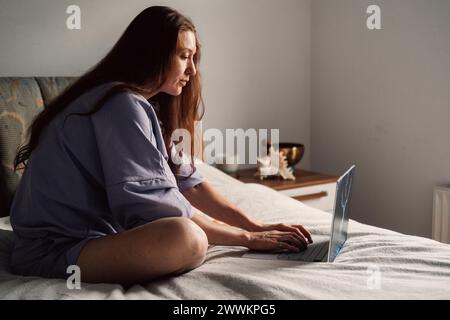 la luce soffusa del mattino illumina una donna concentrata sul proprio computer portatile mentre si siede sul letto, combinando il lavoro Foto Stock