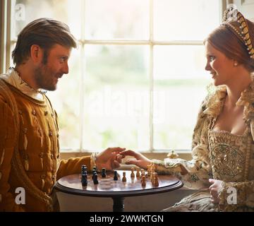 Rinascimentale, coppia e scacchi a casa con tenersi per mano, costume vintage e medievale in un palazzo. Finestra, lusso, letto queen size e sorriso Foto Stock