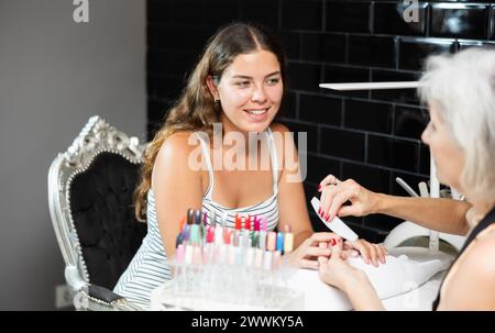 Donna anziana amichevole che lavora nel salone di bellezza come maestro di manicure modellando le unghie di una giovane cliente femminile con una lima per unghie Foto Stock