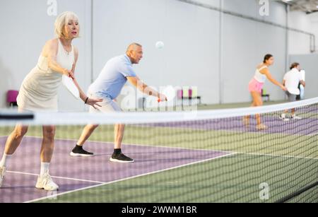 Donna anziana che gioca a double pickleball con un partner maschile al chiuso Foto Stock