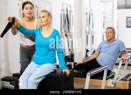 Giovane istruttrice positiva che controlla i movimenti di donna matura che esercita su attrezzature di riformatore Pilates in studio Foto Stock