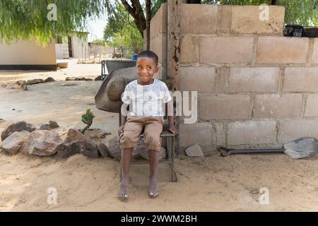 felice bambino del villaggio africano seduto su una sedia nel cortile, casa sullo sfondo Foto Stock