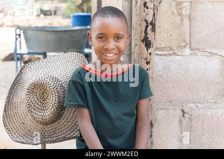 felice villaggio africano bambino seduto nel cortile, casa sullo sfondo Foto Stock