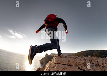 Escursionista attivo con zaino salta su una grande roccia contro il cielo e il mare al tramonto Foto Stock