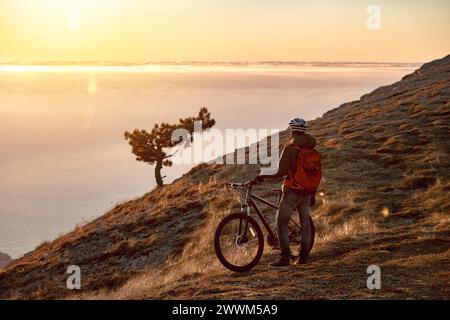 L'atleta sportivo è in piedi con una bici da mtb in cima alla montagna e gode del tramonto o dell'alba Foto Stock