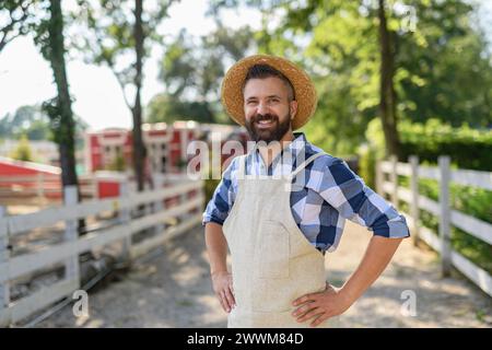 Ritratto di un agricoltore in piedi davanti a una recinzione di legno che guarda la sua fattoria. Uomo con cappello che lavora in un ranch di famiglia. Foto Stock