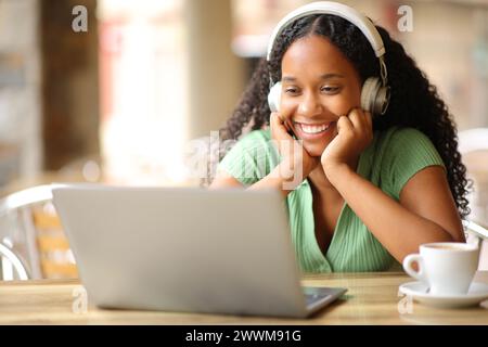 Donna nera felice che guarda i contenuti multimediali utilizzando un computer portatile e le cuffie nella terrazza di un ristorante Foto Stock