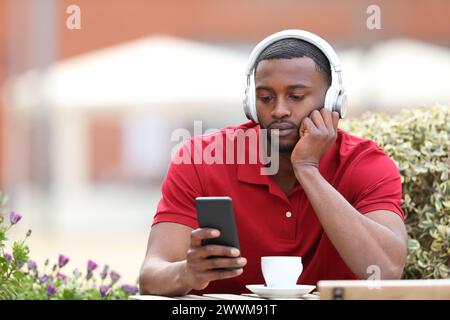 Uomo nero che ascolta la musica al telefono indossando le cuffie in una terrazza del bar Foto Stock