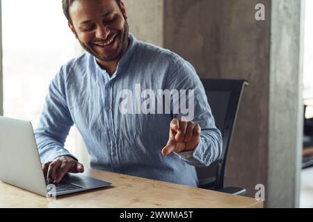 Un uomo che indossa gli occhiali siede su un tavolo di legno, sorride, usando un computer portatile Foto Stock