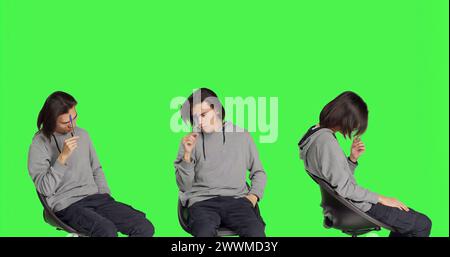 Modello maschile che pensa alla soluzione sullo sfondo verde, sentendosi incerto sul nuovo dilemma. Uomo asiatico seduto su una sedia e che agisce confuso, penoso e premuroso. Foto Stock