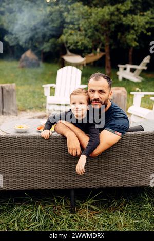 Un uomo seduto su un divano nero con suo figlio nel cortile Foto Stock