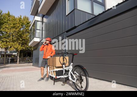 giovane corriere con abbigliamento rosso e casco in bici cargo che arriva alla destinazione di spedizione per consegnare un pacco a un indirizzo della città, parlando con t Foto Stock