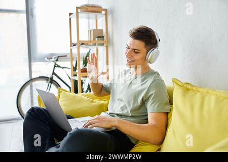 un giovane sorridente con cuffie e laptop sul divano giallo che saluta le riunioni online lateralmente Foto Stock