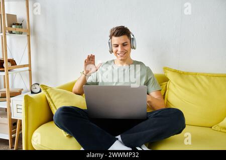 allegro studente con cuffie e laptop sul divano giallo che saluta le riunioni online Foto Stock