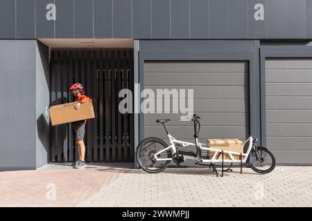 giovane corriere con abbigliamento rosso e casco in bici cargo che arriva alla destinazione di spedizione per consegnare il pacco a un indirizzo della città Foto Stock