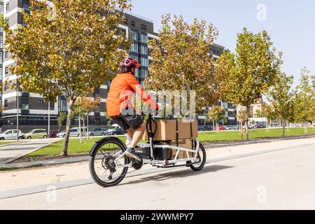 giovane corriere con abbigliamento rosso e casco in bici cargo , percorrendo la pista ciclabile della città per consegnare un pacchetto. Foto Stock