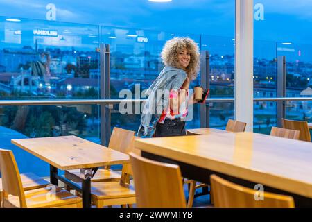 Ritratto di una giovane donna caucasica sorridente con capelli biondi ricci, che lavora in un bar con il suo computer portatile e lo smartphone, che prende e se ne va Foto Stock