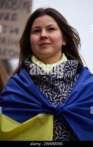 Ritratto di allegra donna Ucraina che indossa una bandiera nazionale dell'Ucraina sulle spalle durante una manifestazione a sostegno delle forze armate. Kiev - 23 marzo 2024 Foto Stock