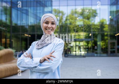 Una donna professionista in un hijab si trova con fiducia e con le braccia incrociate all'esterno di un edificio di uffici contemporaneo, simboleggiando l'empowerment e la diversità sul posto di lavoro. Foto Stock