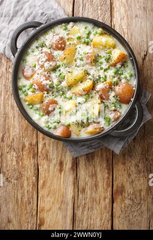 Patate al forno vegetariane e piselli verdi in cremosa salsa al formaggio da vicino in una padella su un tavolo di legno. Vista dall'alto verticale Foto Stock