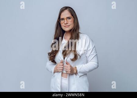 Ritratto in vita di una giovane donna d'affari latina felice, in elegante tuta bianca con occhiali e lunghi capelli marroni, in piedi in posa con una giacca sorridente Foto Stock