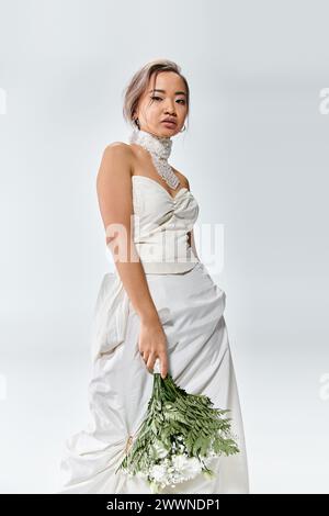 affascinante giovane donna asiatica in elegante abito bianco che posa con bouquet di fiori su sfondo chiaro Foto Stock