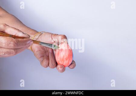 Mano di una donna indiana che regge un uovo di Pasqua e dipinge con un pennello su sfondo bianco Foto Stock