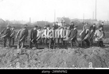 Costruzione di ferrovie. Il gruppo di lavoratori della costruzione ferroviaria tra di loro è visto in uno sforzo di squadra con barre di scavo, che spostano la ferrovia in posizione. anni '1900 Foto Stock