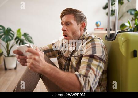 Uomo con la faccia scioccata, seduto sul pavimento con la valigia, che sembra preoccupato e teso sullo schermo del telefono cellulare Foto Stock