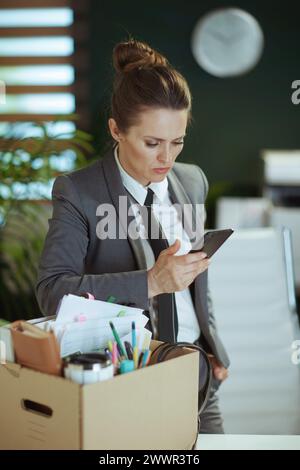 Nuovo lavoro. infelice lavoratrice moderna di mezza età in un moderno ufficio verde in un abito da lavoro grigio con effetti personali in scatola di cartone e smartphone Foto Stock