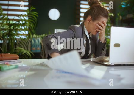 Nuovo lavoro. lavoratrice moderna e triste di mezza età in un moderno ufficio verde con tuta da lavoro grigia e laptop che lancia documenti. Foto Stock
