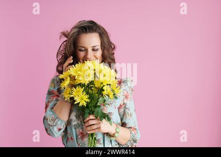 donna alla moda sorridente con lunghi capelli di bruna ondulati con fiori di crisantemi gialli utilizzando uno smartphone su sfondo rosa. Foto Stock