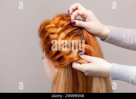 Bellissimo, con una lunga ragazza pelosa dai capelli rossi, parrucchiere intreccia una treccia, primo piano in un salone di bellezza. Cura professionale dei capelli e creazione di acconciature. Foto Stock