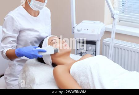 Il medico-cosmetologo rende l'apparecchio una procedura di pulizia ecografica della pelle del viso di una bella, giovane donna in un salone di bellezza. Cosmo Foto Stock