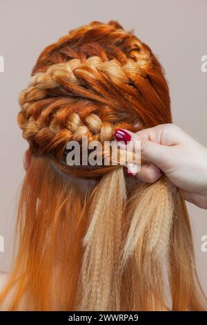 Bellissimo, con una lunga ragazza pelosa dai capelli rossi, parrucchiere intreccia una treccia, primo piano in un salone di bellezza. Cura professionale dei capelli e creazione di acconciature. Foto Stock