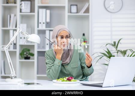 Vista frontale di una donna turbata seduta alla scrivania con un computer portatile moderno e forchetta per mangiare insalata. Una donna araba frustrata si sente troppo a mangiare e guarda il piatto con disgusto in ufficio. Foto Stock