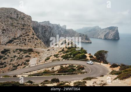 Piccola auto bianca solitaria che guida su una strada di montagna asfaltata curva a serpentina vicino al faro di Cap de Formentor con un bellissimo paesaggio marino con costa rocciosa Foto Stock