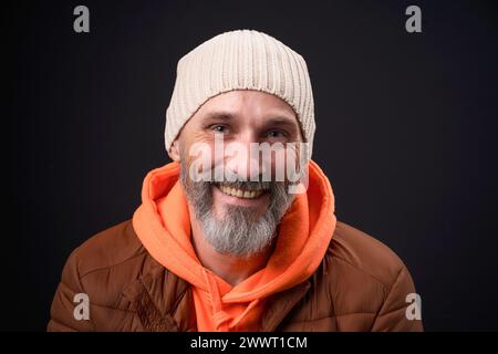 Un uomo che indossa una giacca marrone e una felpa con cappuccio arancione sorride. Ha un cappello bianco in maglia sulla testa Foto Stock