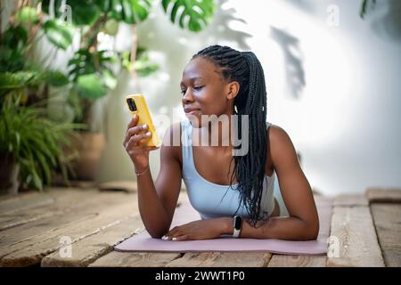 Una giovane donna afroamericana che si rilassa sul tappetino per il fitness con lo smartphone dopo l'allenamento. Foto Stock