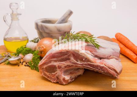 Spalla fresca di agnello crudo su un tagliere di legno con erbe fresche, sale e grani di pepe , olio d'oliva, cipolle, patate, mortaio e pestello Foto Stock