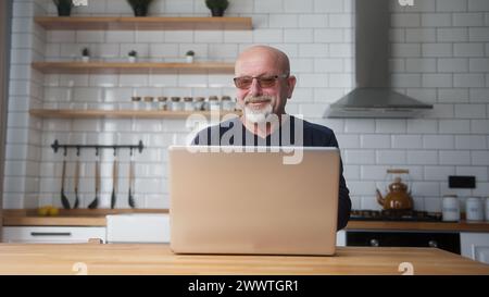 Un uomo sorridente e maturo in vetro si siede in cucina utilizzando il laptop, trascorri il tempo libero su Internet, goditi la comunicazione sui social media, riposati a casa. Wireless Foto Stock