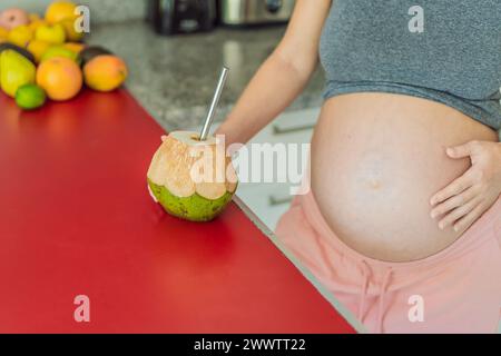 Dissetandosi dalla sua sete di gravidanza con una scelta rinfrescante, una donna incinta beve gioiosamente l'acqua di cocco da una noce di cocco in cucina, abbracciando Foto Stock